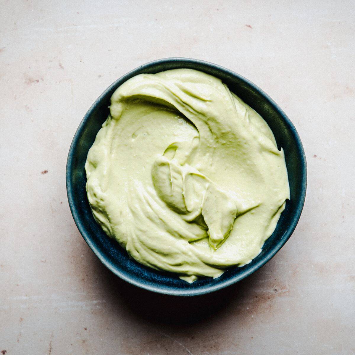 A small dark bowl filled with smooth, creamy light green avocado crema sits on a light-colored surface.