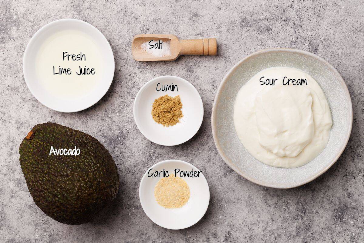 Top-down view of ingredients for avocado crema: a whole avocado, a bowl of sour cream, a bowl of fresh lime juice, a small bowl of cumin, a small bowl of garlic powder, and a wooden spoon with salt.