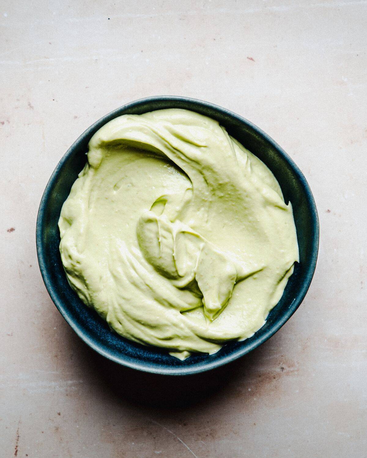Avocado Crema in a dark bowl, featuring a creamy green texture and a light colored background.