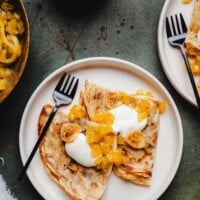 A plate with folded banana crepes topped with whipped cream and caramelized bananas, served with a black fork. Another plate and a pan with more crepes are partially visible. The text reads "Banana Crepes" and "pinchandswirl.com.