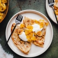 Two folded banana crepes on a white plate topped with cream, caramelized banana slices, and pineapple marmalade. A black fork rests on the plate, and part of another plate with similar food is visible in the corner.
