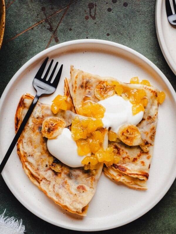 Two folded banana crepes on a white plate topped with cream, caramelized banana slices, and pineapple marmalade. A black fork rests on the plate, and part of another plate with similar food is visible in the corner.