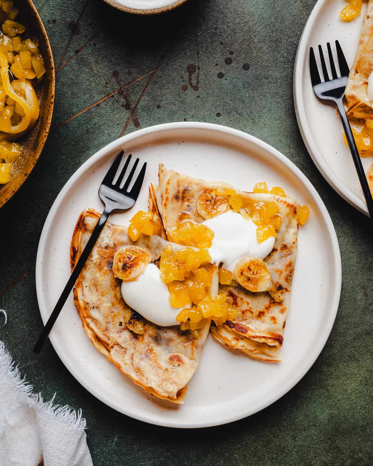 A plate with folded banana crepes topped with sweetened sour cream and caramelized bananas and pineapple marmalade. A black fork rests on the plate, and another partially visible plate is nearby on a dark green surface.