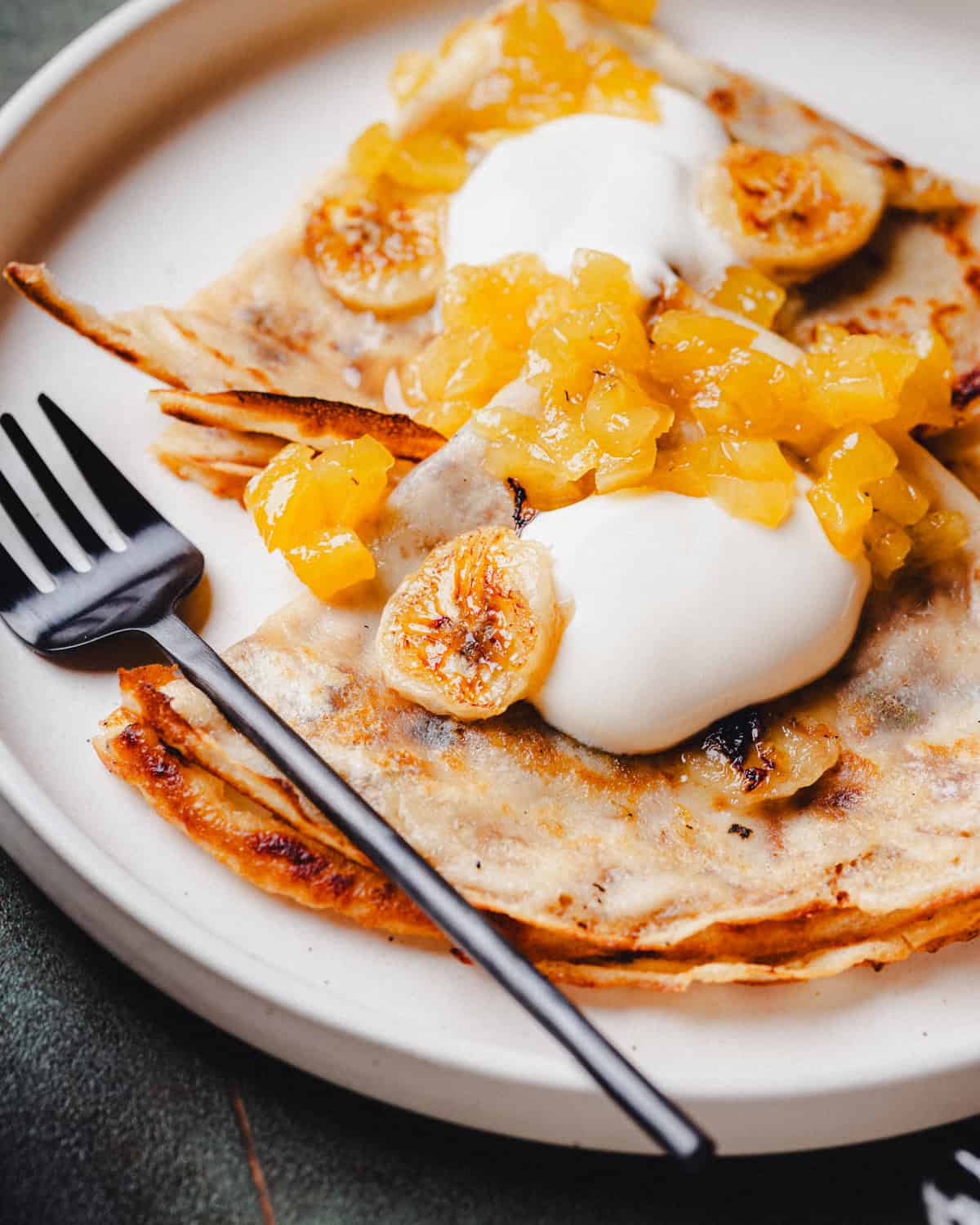 A plate with folded banana crepes topped with caramelized banana slices, pineapple chunks, and dollops of cream, next to a black fork on a white plate.
