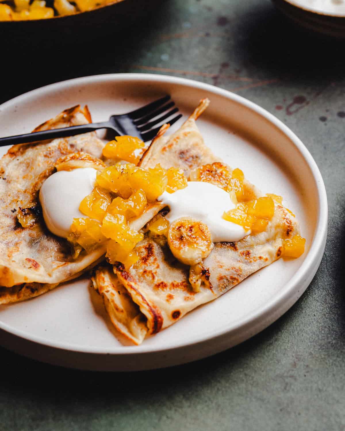 Two folded banana crepes on a white plate, topped with dollops of cream and a generous spoonful of diced yellow fruit compote, with a black fork placed on top.