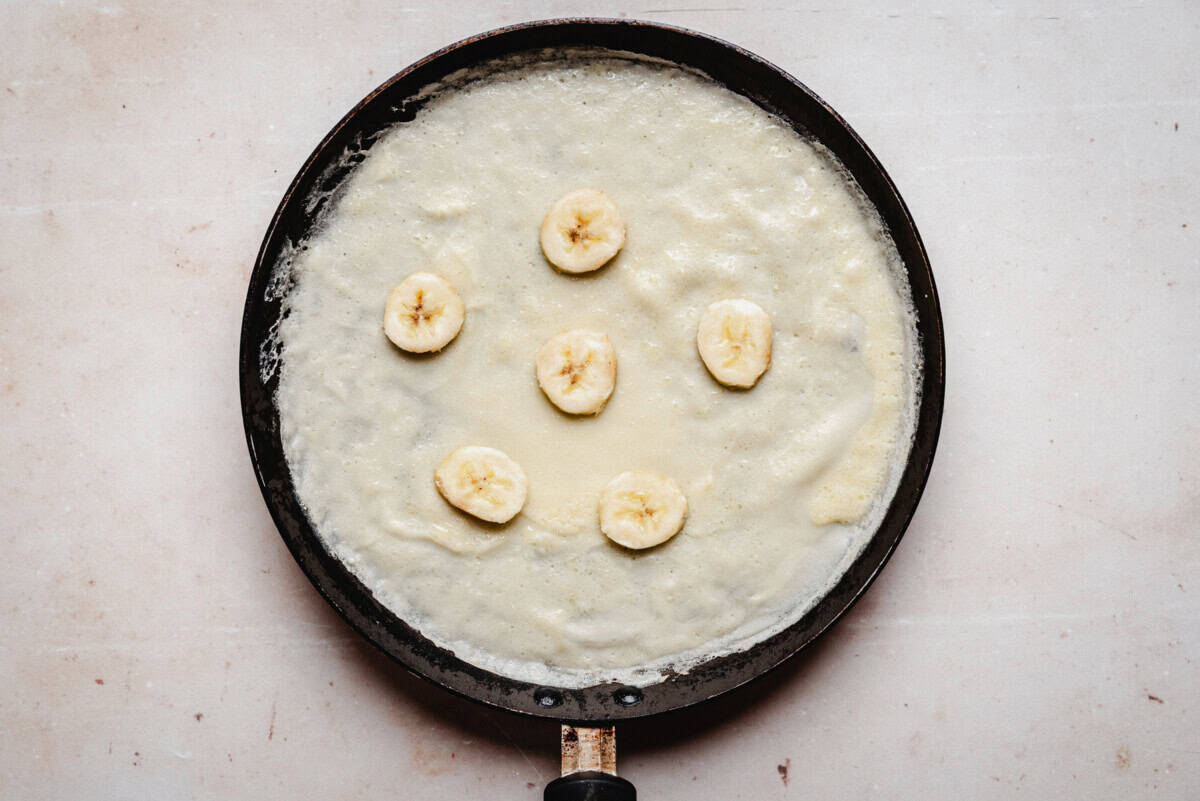 Banana crepe cooking in a frying pan on stovetop, topped with banana slices.