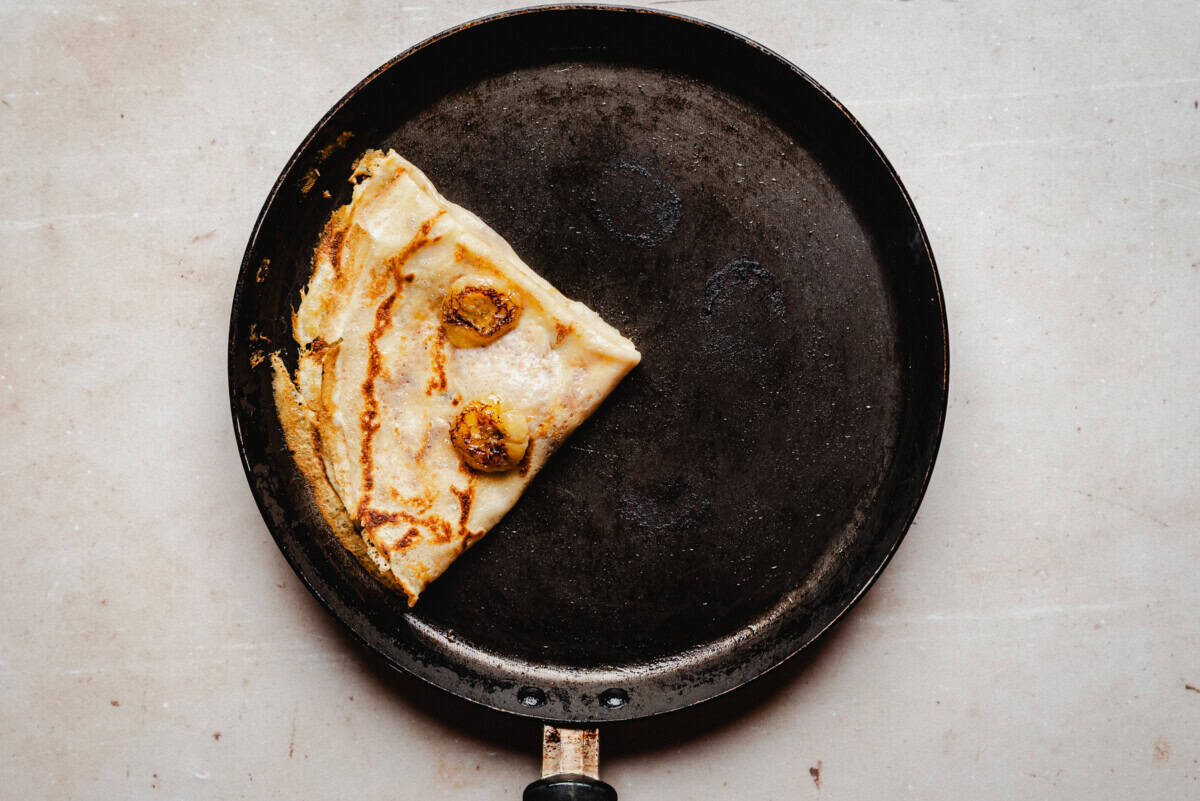 Banana crepe folded and lightly browned, topped with banana slices in a black frying pan.