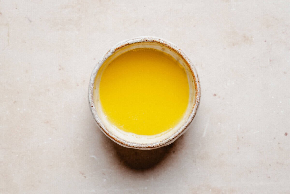 Melted butter in a rustic ceramic bowl, bright yellow and viewed from above on a beige surface.