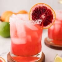 A glass of blood orange margarita with ice, a salted rim, and a slice of blood orange garnish, set on a wooden coaster with lime wedges and fruit blurred in the background.