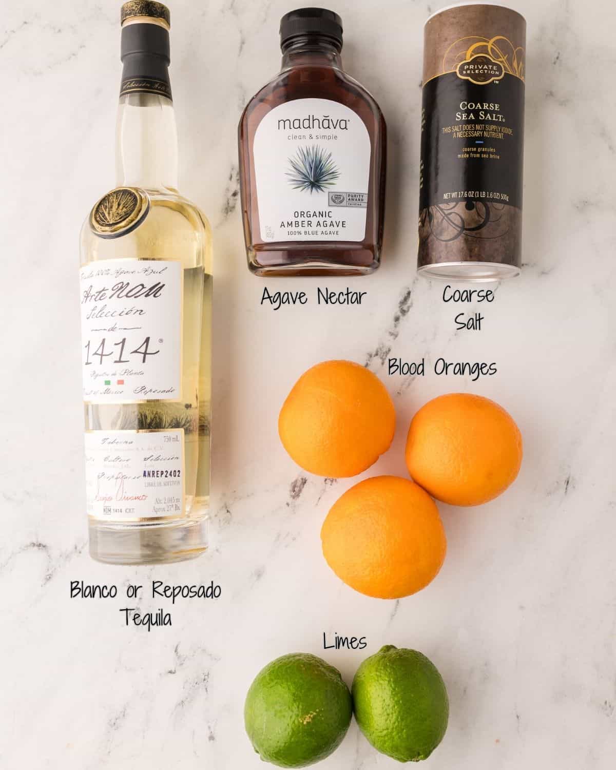 Top-down view of blood orange margarita ingredients on a marble surface: a bottle of tequila, a bottle of agave nectar, a container of coarse sea salt, three blood oranges, and two limes, each labeled with handwritten text.