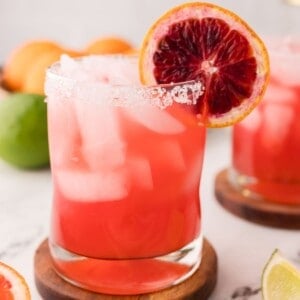 A glass of Blood Orange Margarita with ice, a salted rim, and a slice of blood orange garnish, set on a wooden coaster with citrus fruits blurred in the background.