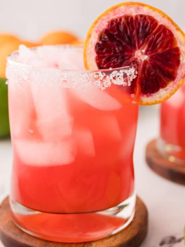 A glass of Blood Orange Margarita with ice, a salted rim, and a slice of blood orange garnish, set on a wooden coaster with citrus fruits blurred in the background.