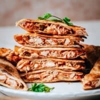 A stack of golden-brown chicken tinga quesadillas filled with shredded chicken and salsa, garnished with cilantro, on a white plate with the website name pinchandswirl.com at the bottom.