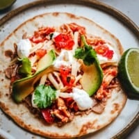 A tostada topped with shredded chicken, sliced avocado, cheese, chopped tomatoes, cilantro, and dollops of sour cream, served with lime wedges on a rustic plate.
