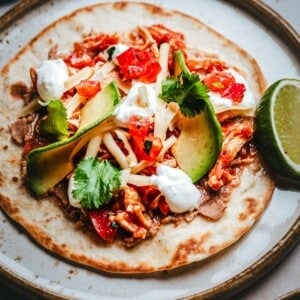 A close-up of a chicken tinga tostada topped with shredded chicken, sliced avocado, cheese, chopped tomatoes, fresh cilantro, and dollops of sour cream, served with a lime wedge on the side.
