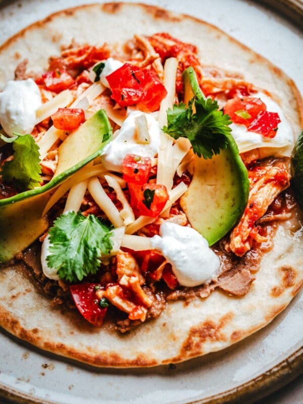 A close-up of a chicken tinga tostada topped with shredded chicken, sliced avocado, cheese, chopped tomatoes, fresh cilantro, and dollops of sour cream, served with a lime wedge on the side.