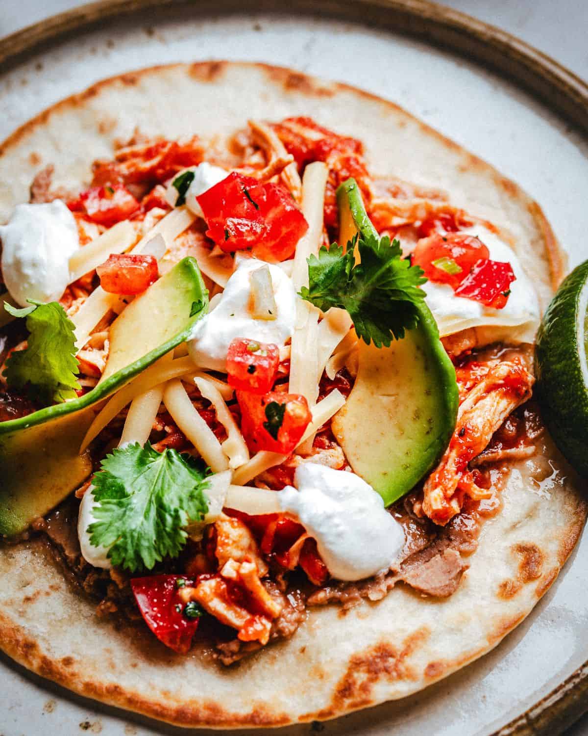 A close-up of a tostada topped with chicken tinga, sliced avocado, shredded cheese, diced tomatoes, cilantro, refried beans, and dollops of sour cream, served with a lime wedge on a plate.