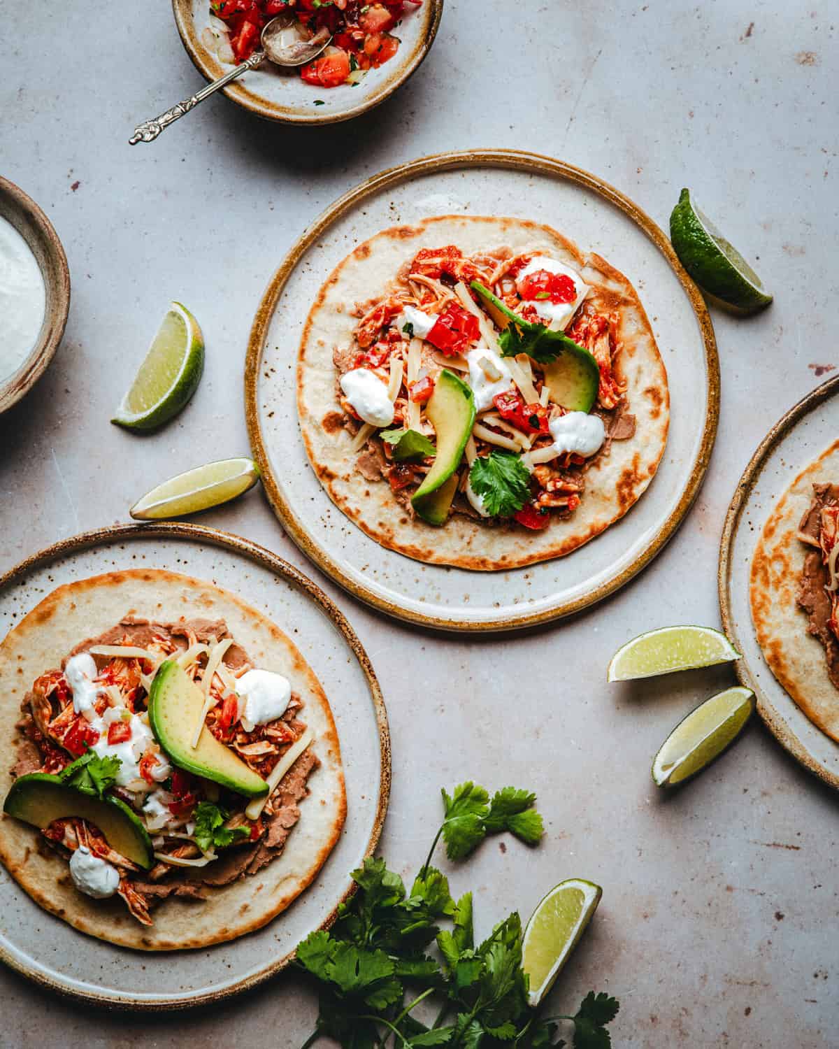 Three plates with chicken tinga tostadas topped with shredded meat, avocado, cheese, salsa, cilantro, and sour cream, surrounded by lime wedges and fresh cilantro on a light table.