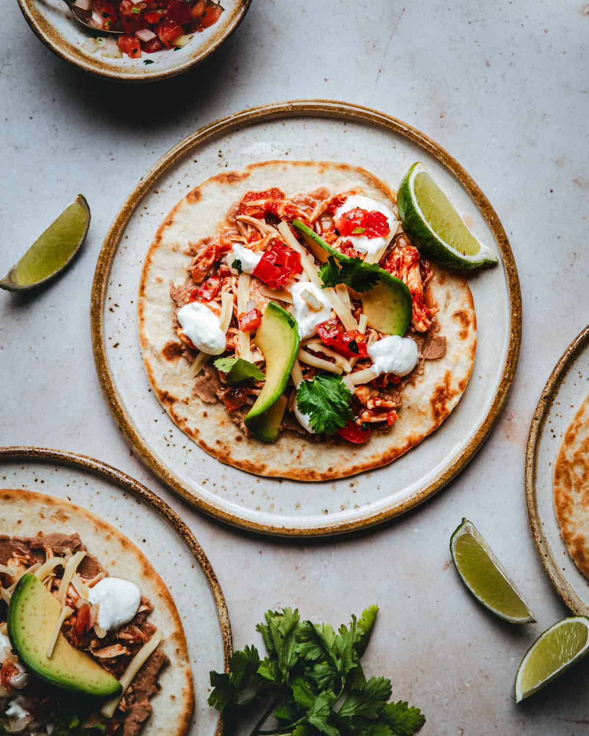 A plate with a tortilla topped with shredded meat, avocado slices, cheese, tomato salsa, cilantro, and sour cream, garnished with lime wedges, surrounded by similar plates and a bowl of salsa.