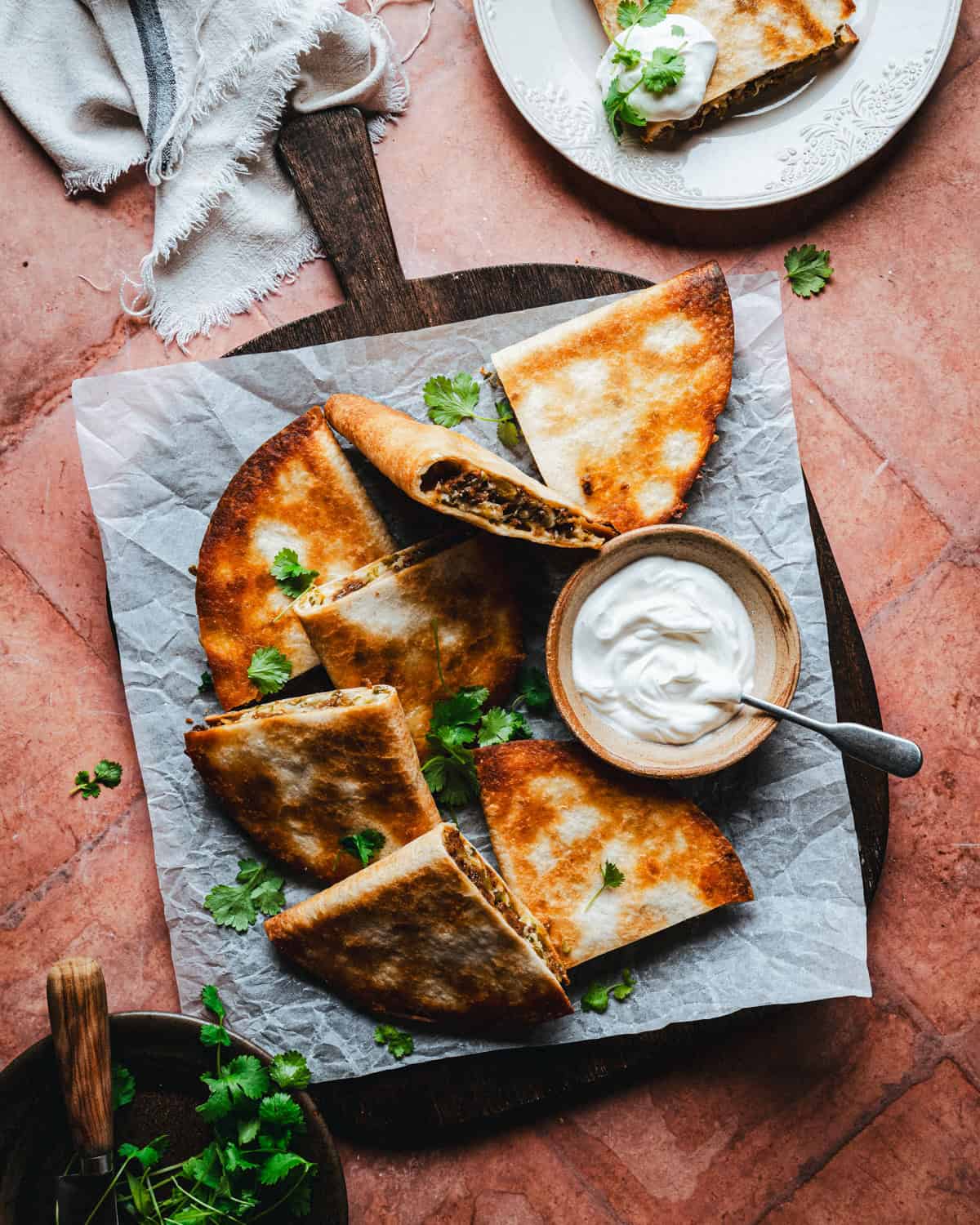 Sliced chorizo quesadillas on parchment paper atop a wooden board, garnished with cilantro, with a bowl of sour cream. A partially visible plate with more quesadilla and sour cream is in the background.