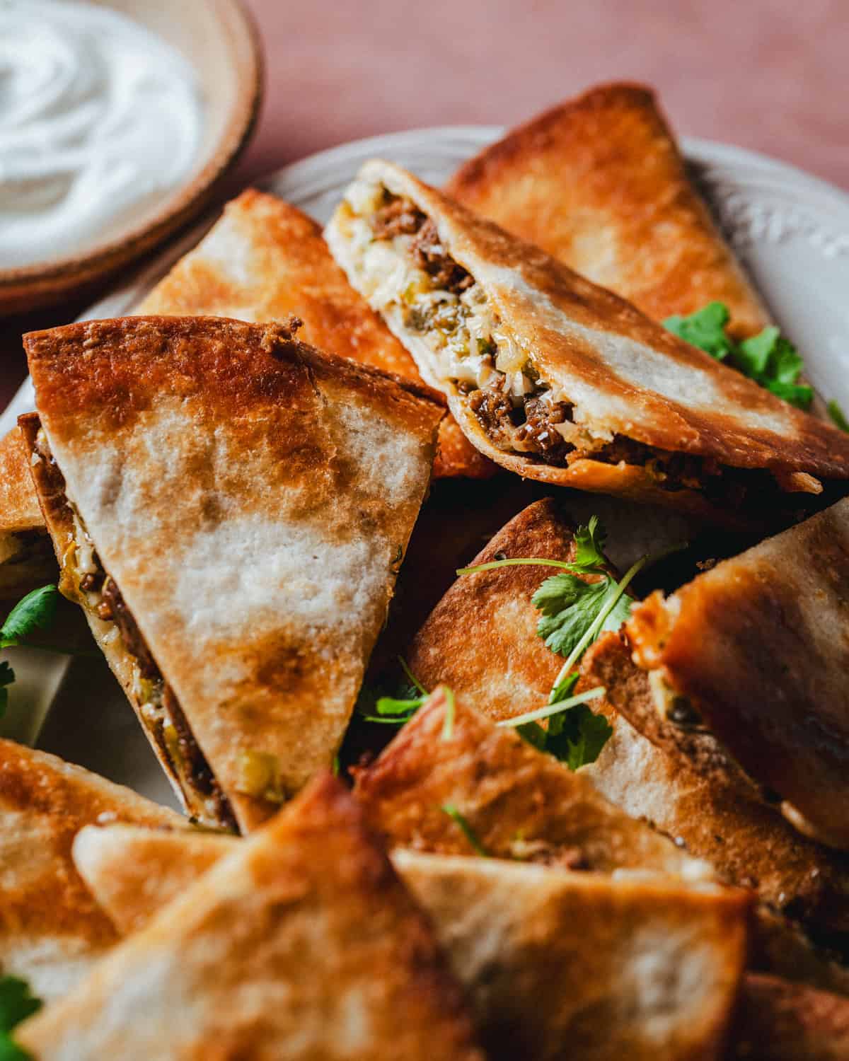 Golden, crispy chorizo quesadilla wedges filled with seasoned ground meat and melted cheese are arranged on a plate, garnished with fresh cilantro, with a bowl of sour cream visible in the background.