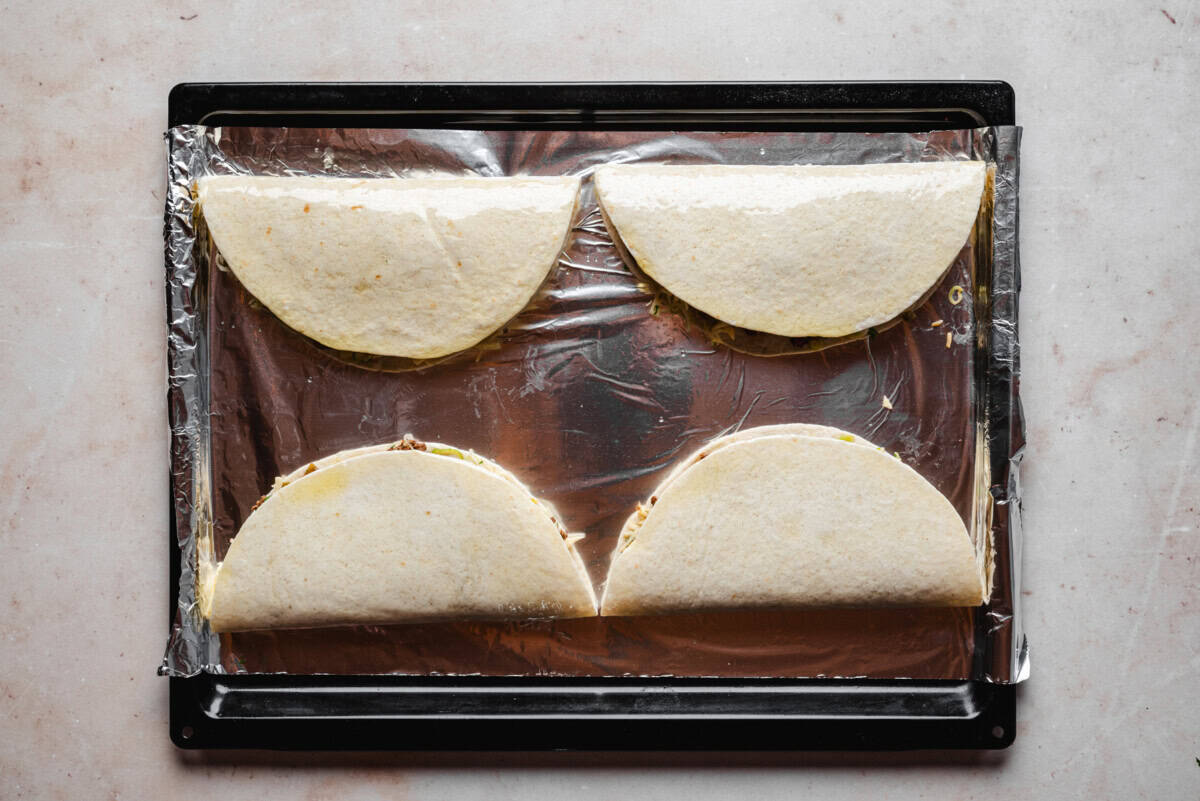 Three folded tortillas filled with ingredients are placed on a baking sheet lined with plastic wrap, ready to be baked or cooked. The tortillas are evenly spaced on the tray.