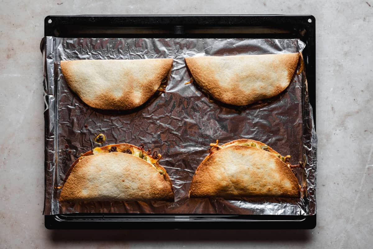 A baking tray lined with foil holds four baked, folded tortillas filled with melted cheese and vegetables, two of which have some filling visible. The tortillas are slightly browned on top.