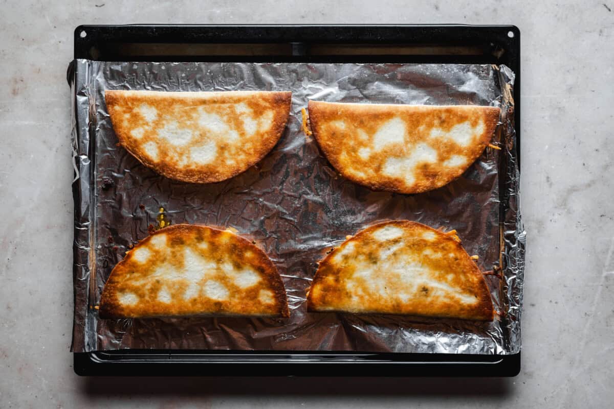 Four folded, golden-brown quesadillas on a foil-lined baking sheet, viewed from above, with some melted cheese spots visible on the tortillas.