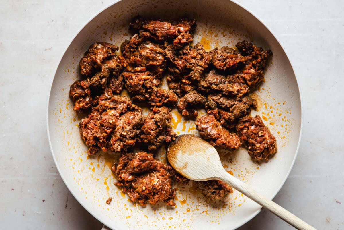 Cooking ground chorizo in a pan, with a wooden spoon resting inside. The meat is browned and slightly oily, and the pan is on a light-colored surface.