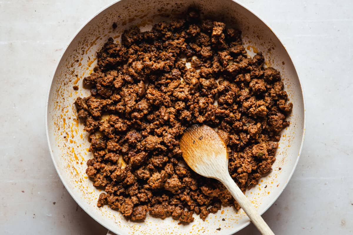 A skillet filled with cooked, crumbled chorizo, with a wooden spoon resting on the side, on a light-colored surface.