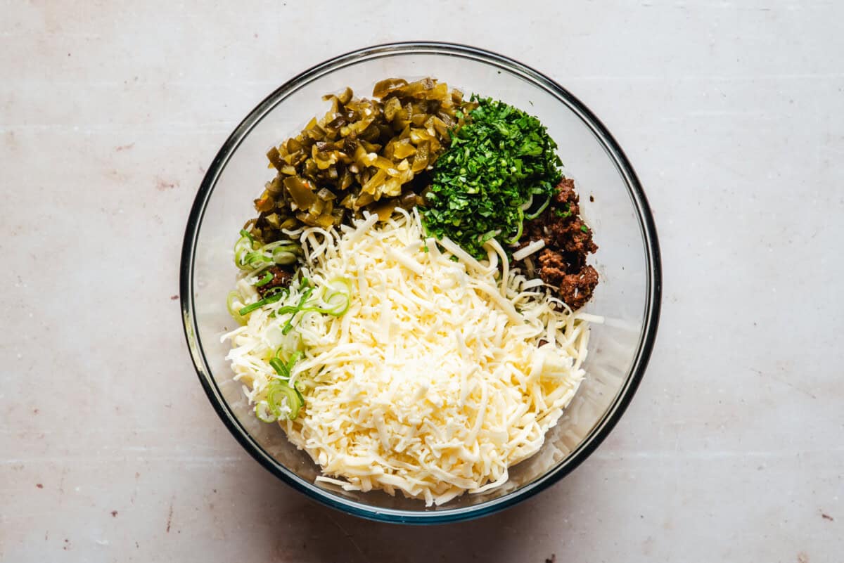 A glass bowl containing shredded cheese, chopped green herbs, sliced green onions, diced green peppers, and crumbly chorizo, all arranged separately on a light surface.