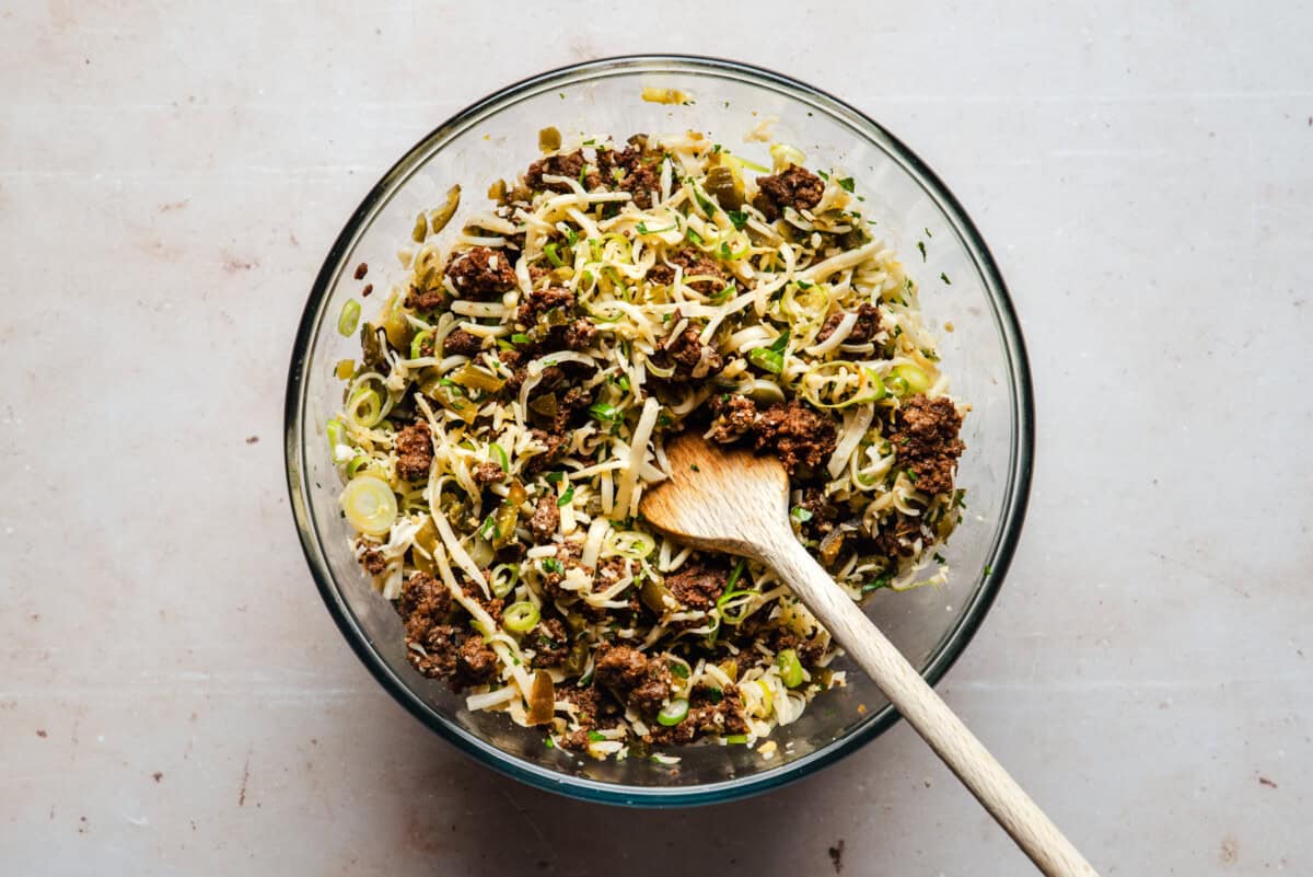 A glass bowl filled with a mixture of cooked ground chorizo, shredded cheese, sliced green onions, and seeds, with a wooden spoon resting inside the bowl, on a light-colored surface.