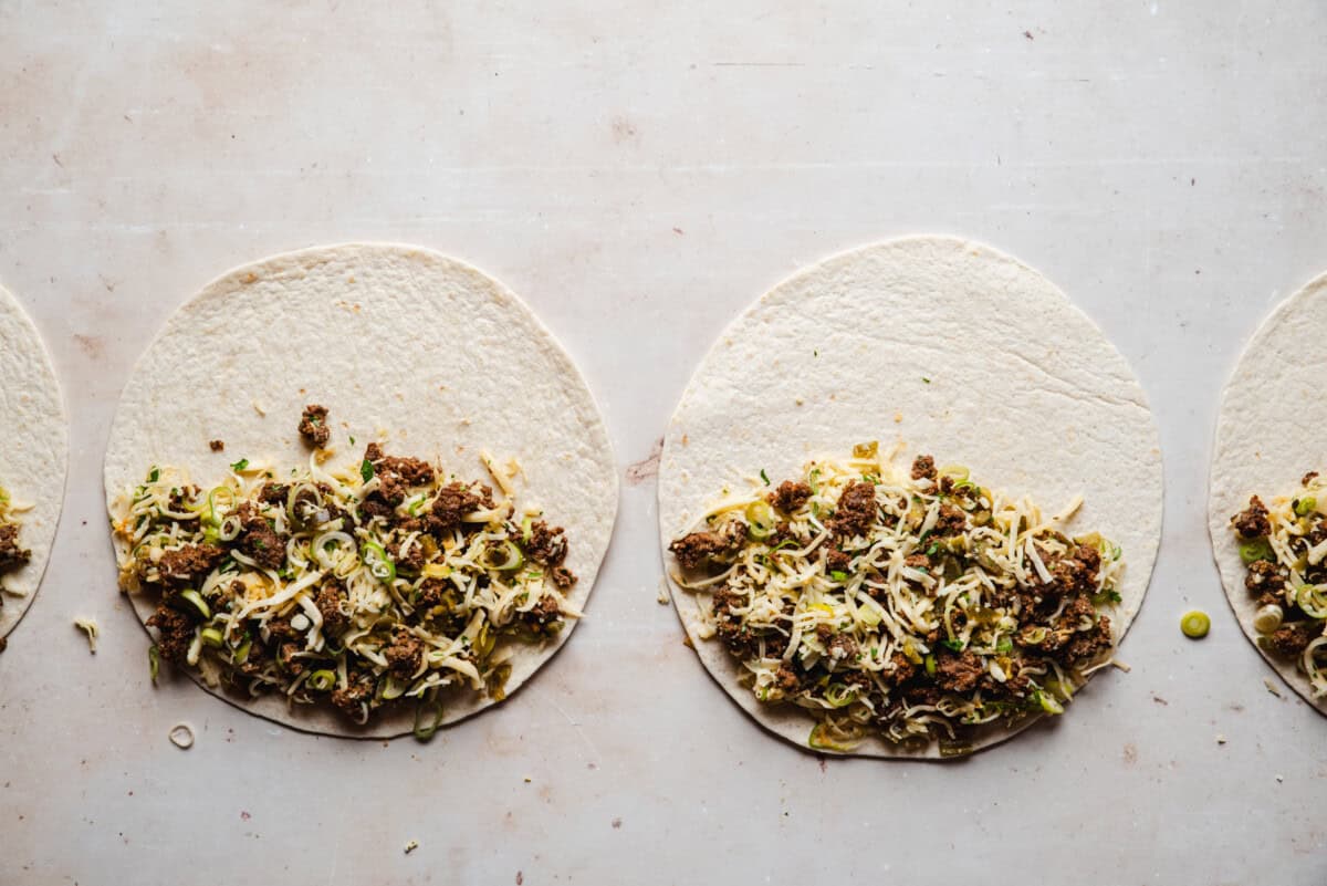 Two flour tortillas on a light surface, each topped with a mixture of cooked and crumbled chorizo, shredded cheese, and sliced green onions, ready to be folded or rolled.