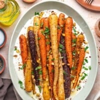 An oval platter of colorful honey roasted carrots garnished with chopped herbs and nuts, served on a bed of creamy sauce. The plate is surrounded by napkins, dishes, and a bottle of olive oil.