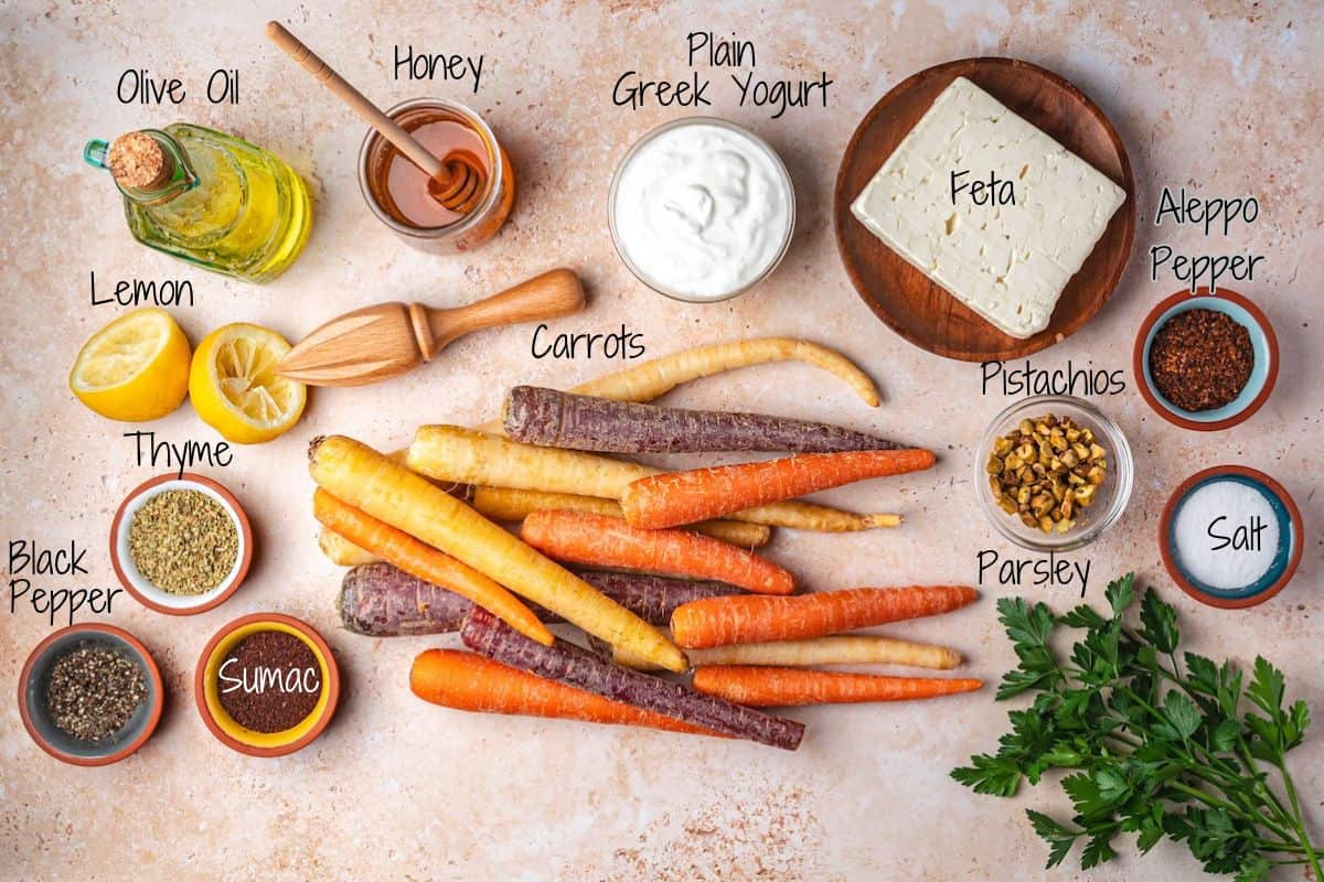 Rainbow carrots surrounded by small bowls of spices, herbs, salt, pistachios, feta cheese, Greek yogurt, olive oil, honey, and lemon halves on a light surface, with each ingredient labeled.