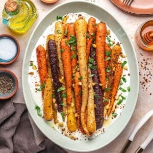 A platter of roasted rainbow carrots on a bed of creamy white sauce, garnished with chopped herbs and nuts, surrounded by bowls of salt, pepper, honey, and a bottle of olive oil.