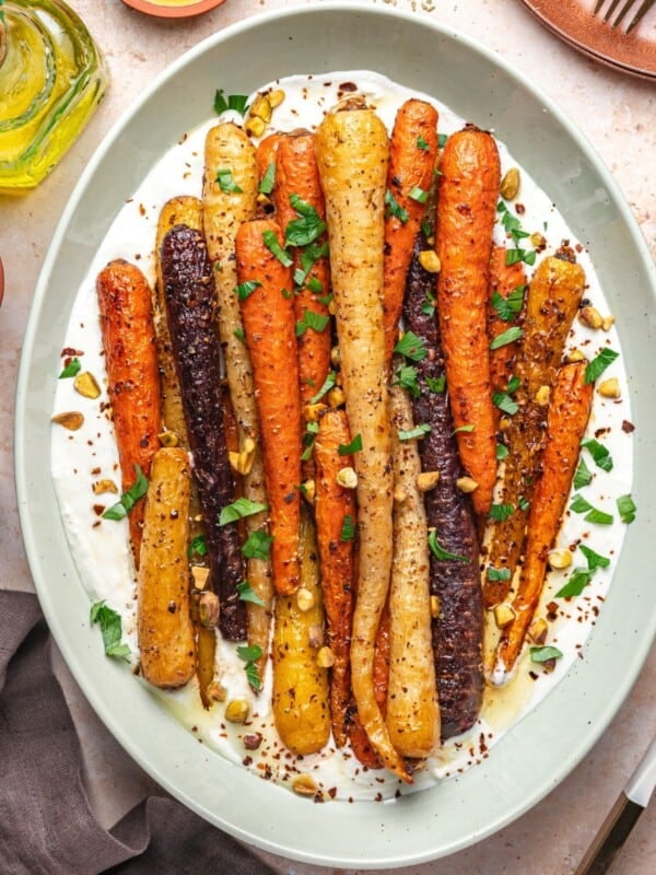 A platter of roasted rainbow carrots on a bed of creamy white sauce, garnished with chopped herbs and nuts, surrounded by bowls of salt, pepper, honey, and a bottle of olive oil.