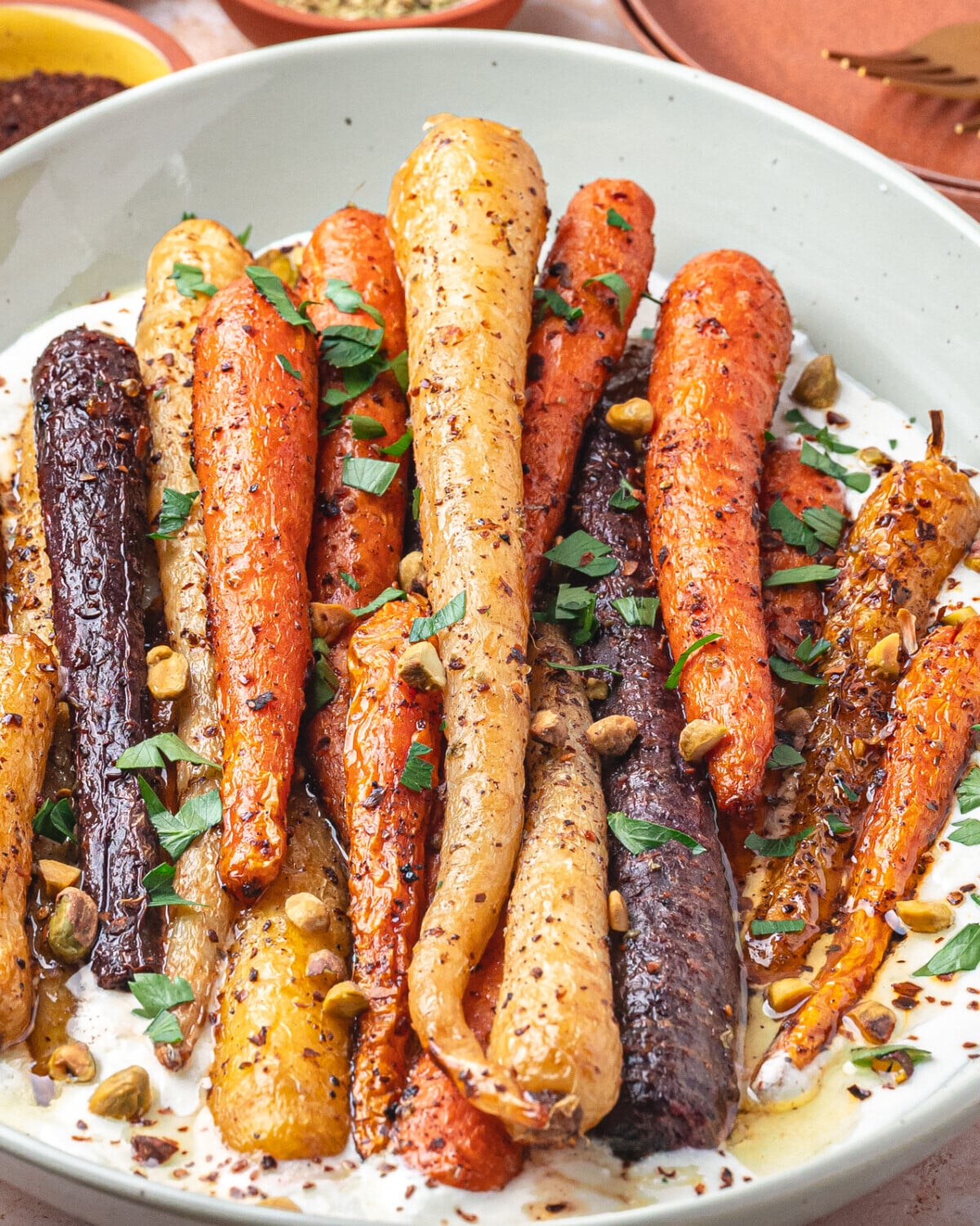 A bowl of honey roasted multicolored carrots garnished with chopped herbs and nuts, served on a creamy white sauce, with small bowls of spices in the background.