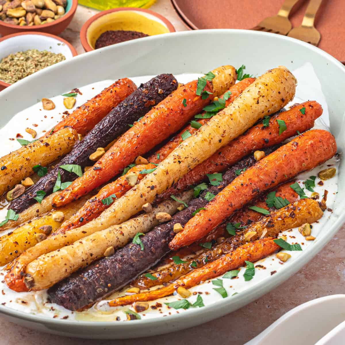 A dish of colorful honey roasted carrots garnished with chopped herbs and nuts, served on a bed of creamy sauce in a white oval bowl, with spices and utensils in the background.