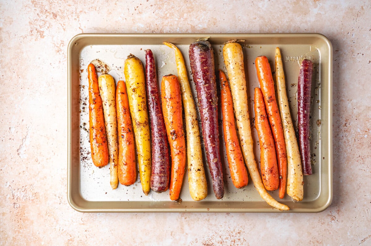A baking sheet with colorful carrots, including orange, yellow, and purple, seasoned with spices on a light tan surface.