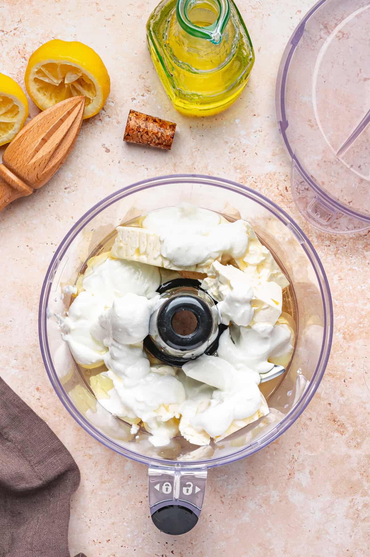 A food processor bowl filled with feta, Greek yogurt sits on a counter next to a bottle of olive oil, a lemon squeezer, half a squeezed lemon, and a cork.