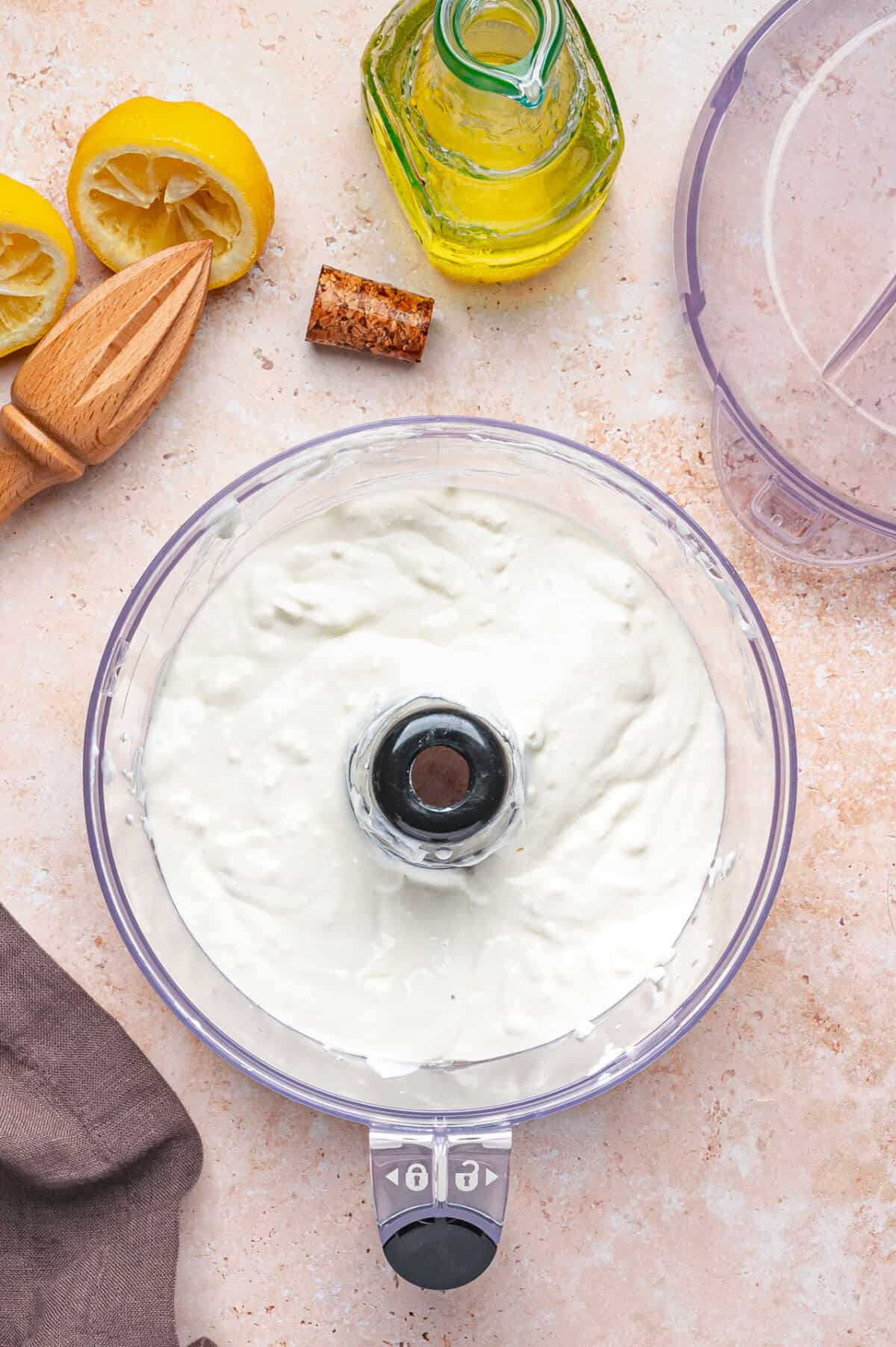 A food processor bowl filled with whipped feta sits on a counter, surrounded by a halved lemon, cork, bottle of olive oil, wooden citrus reamer, and a brown napkin.