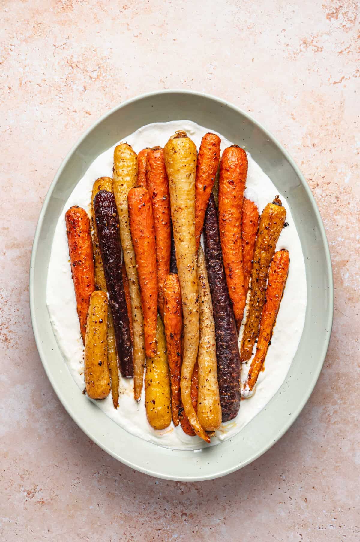 A plate of roasted rainbow carrots, including orange, yellow, and purple, arranged on a layer of creamy white sauce in a light green dish.