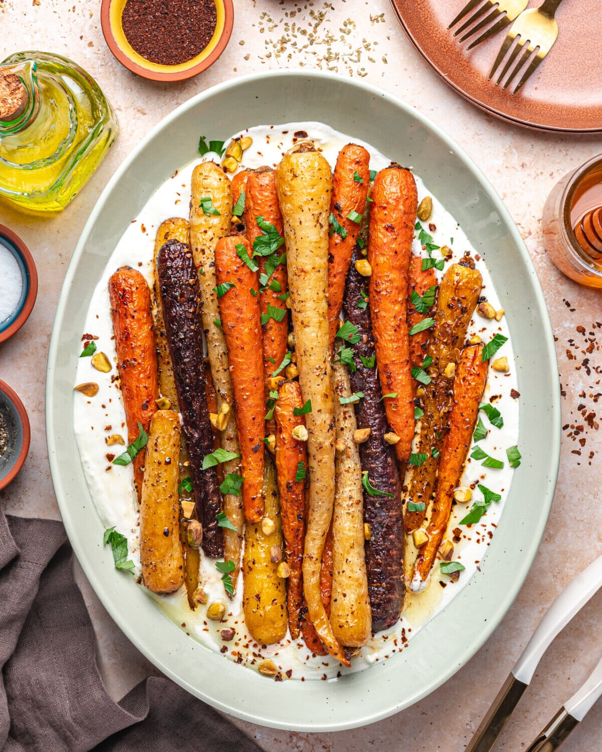 A platter of roasted colorful honey roasted carrots on a bed of whipped feta, garnished with chopped herbs and nuts. Surrounding the dish are small bowls of spices, olive oil, empty plates, and utensils.