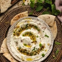 A bowl of creamy labneh dip garnished with herbs and pistachios is surrounded by pita bread pieces on a woven tray; fresh mint leaves are scattered around.