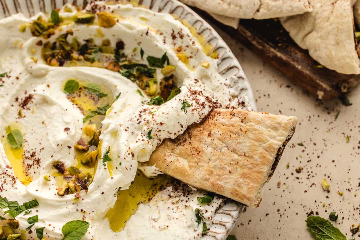 A close-up of creamy labneh dip garnished with chopped pistachios, herbs, olive oil, and spices, with a piece of pita bread being dipped into it. Other pita pieces are visible in the background.