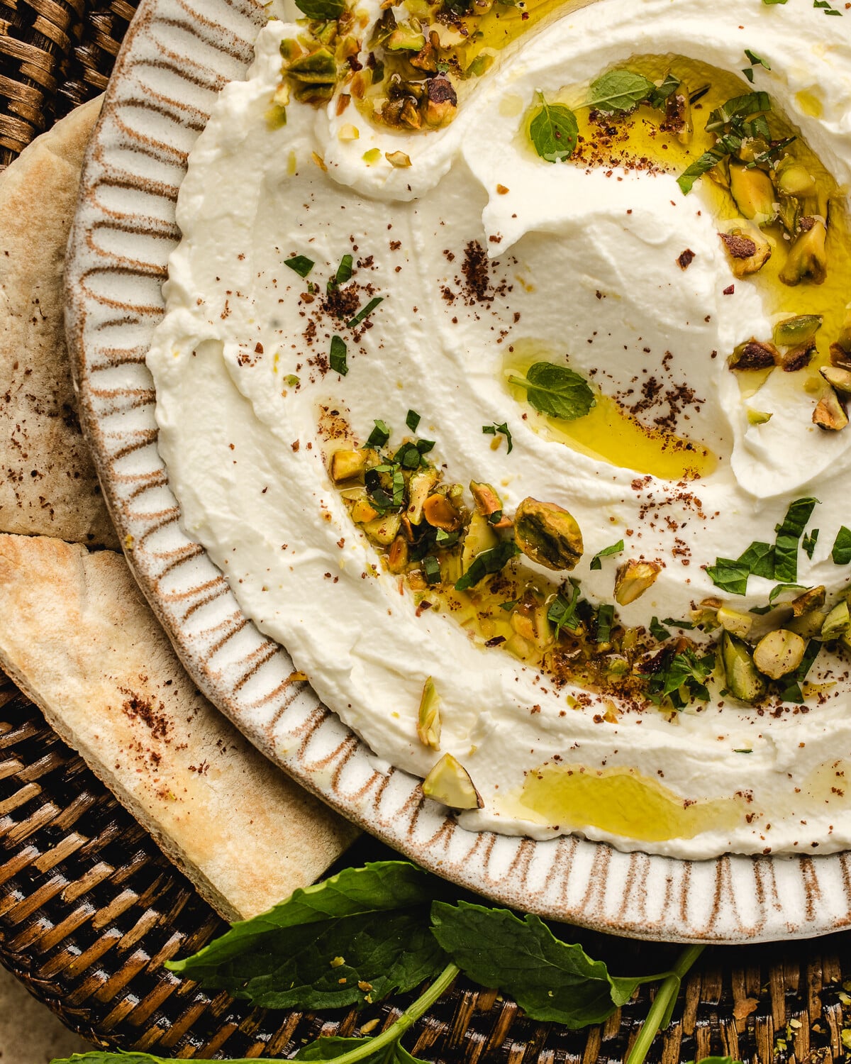 A close-up of creamy labneh dip topped with olive oil, chopped pistachios, herbs, and sumac, served on a white plate with pieces of flatbread and fresh mint leaves on the side.