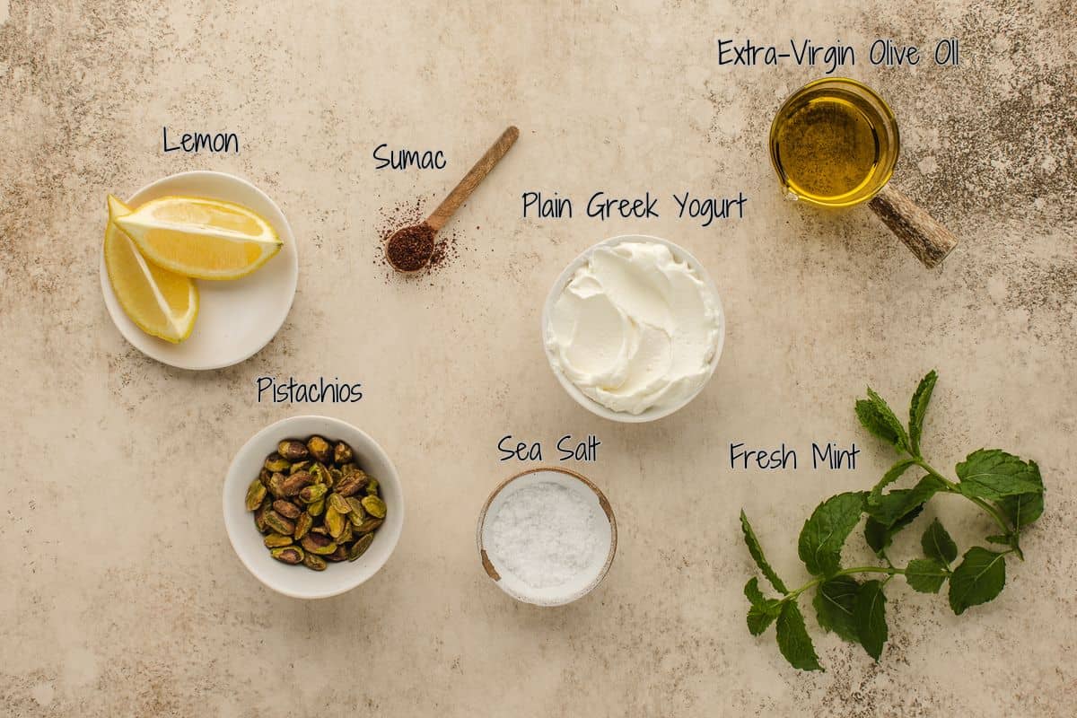Overhead view of labneh dip ingredients labeled: lemon wedges, sumac powder, plain Greek yogurt, extra-virgin olive oil, pistachios, sea salt, and fresh mint on a textured surface.