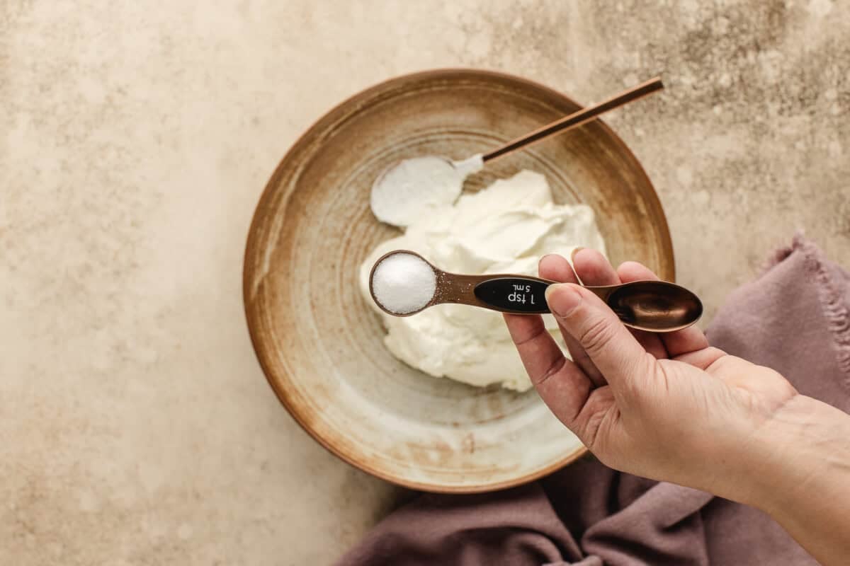 A hand holds a measuring spoon labeled "1 tsp salt" above a brown bowl containing greek yogurt, with a spoon resting inside the bowl on a beige surface.