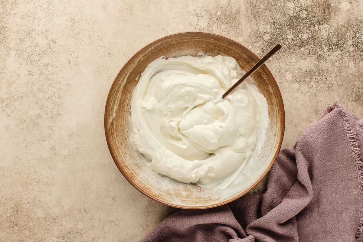 A bowl of creamy greek yogurt mixed with salt with a spoon that rests on a beige surface next to a folded mauve cloth.